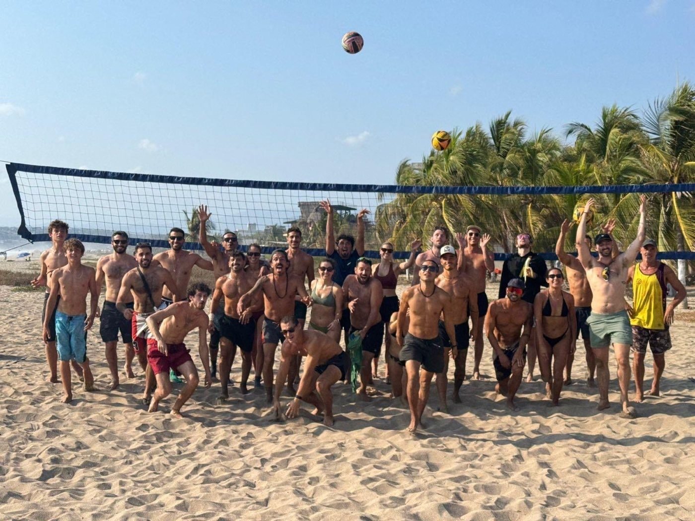 A large group of players at the net on the beach in Puerto Escondido, arms raised, ball in the air.